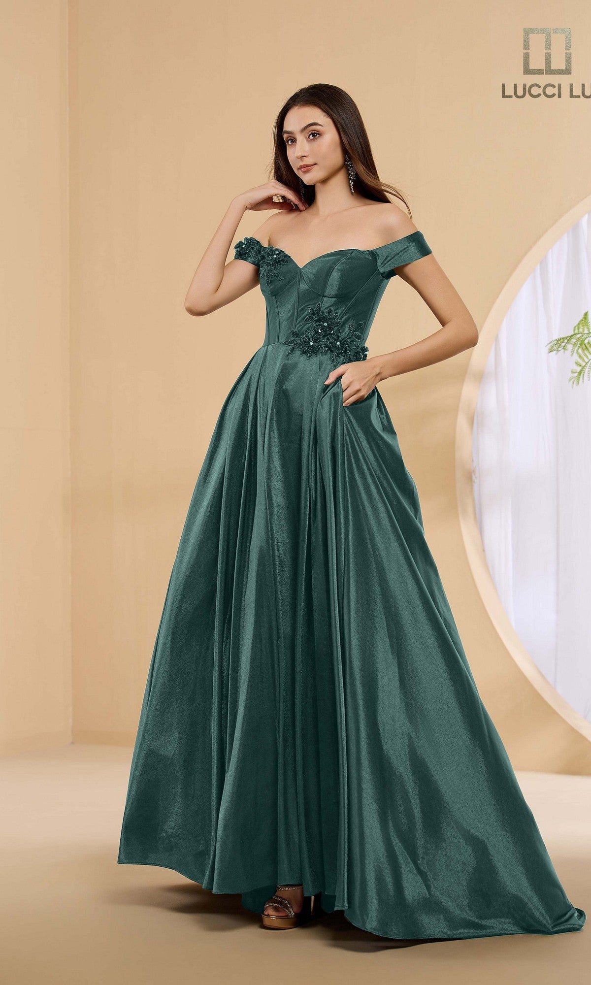 A woman poses indoors in the Lucci Lu 1472 long prom dress, an off-the-shoulder A-line gown in hunter green with floral details and pockets.